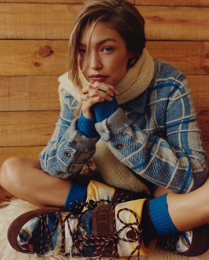 Gigi Hadid in a blue patterned jacket and cream scarf seated against a wooden wall for Guest In Residence Winter 2025