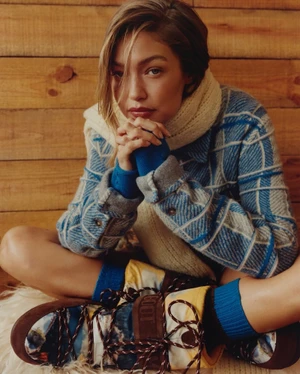 Gigi Hadid in a blue patterned jacket and cream scarf seated against a wooden wall for Guest In Residence Winter 2025
