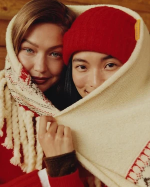 Gigi Hadid and another model wrapped in a cream patterned blanket and wearing a red beanie for Guest In Residence.