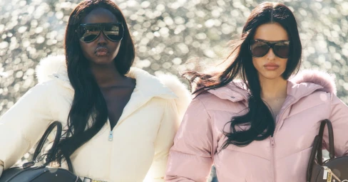 Adut Akech and Amelia Gray wearing winter puffer jackets and sunglasses while holding black handbags.