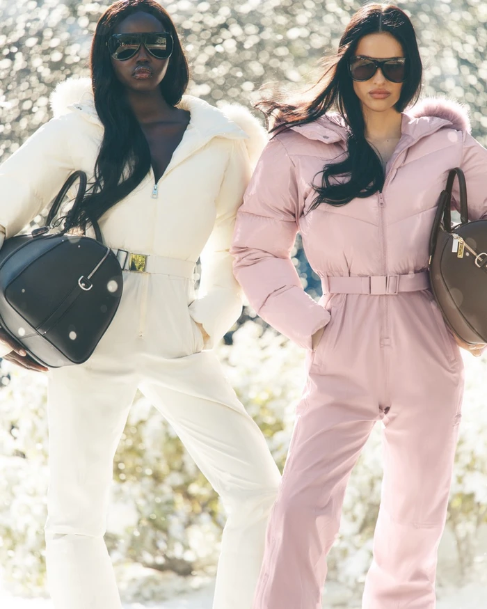 Adut Akech and Amelia Gray wearing winter ski jumpsuits and holding handbags in an outdoor winter setting