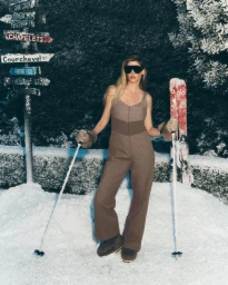 Model Lara Stone in a brown ski suit with sunglasses holding ski poles standing in snow for Alo Winter 2025.