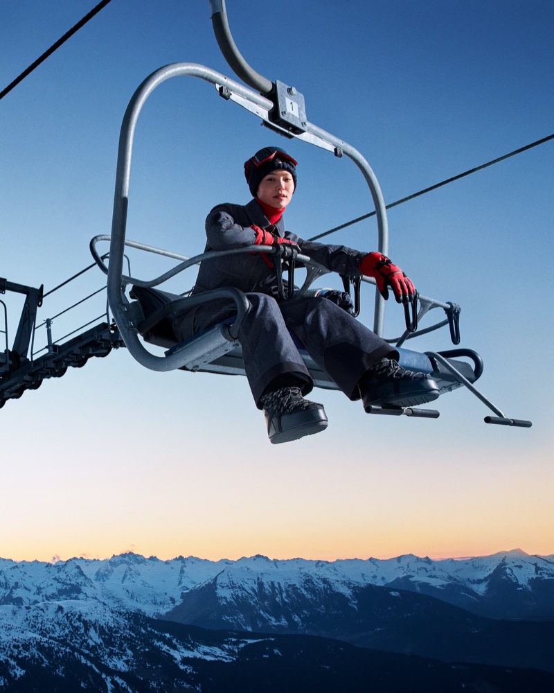Snowboarder Chloe Kim sitting on a ski lift chair wearing grey winter outerwear with red gloves above snow-capped mountain peaks.