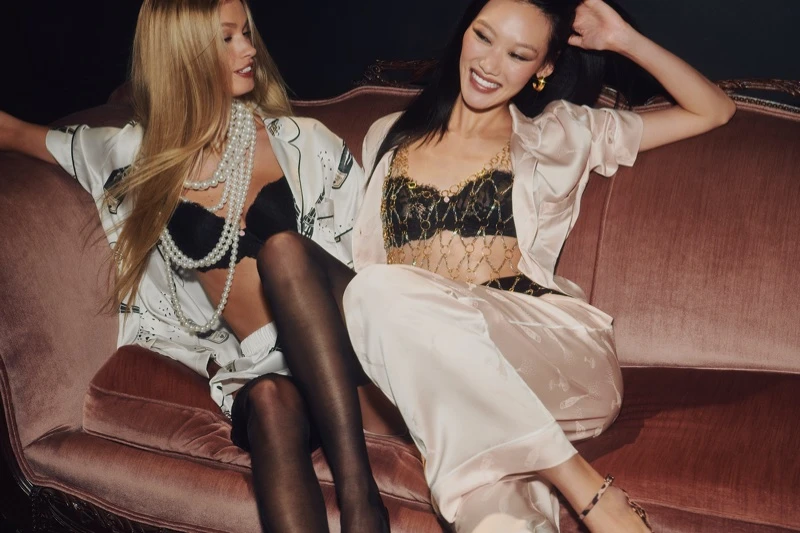 Two female models seated together on a plush pink velvet sofa wearing lingerie and silk pajama sets for Victoria's Secret.