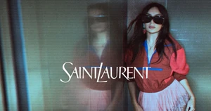 Woman in oversized red jacket and pink pleated skirt wearing sunglasses next to a glass reflection for Yves Saint Laurent.