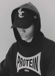 Kevin Nyavedji wearing a black 032c Protein hoodie and branded baseball cap looking down in a monochrome studio portrait.