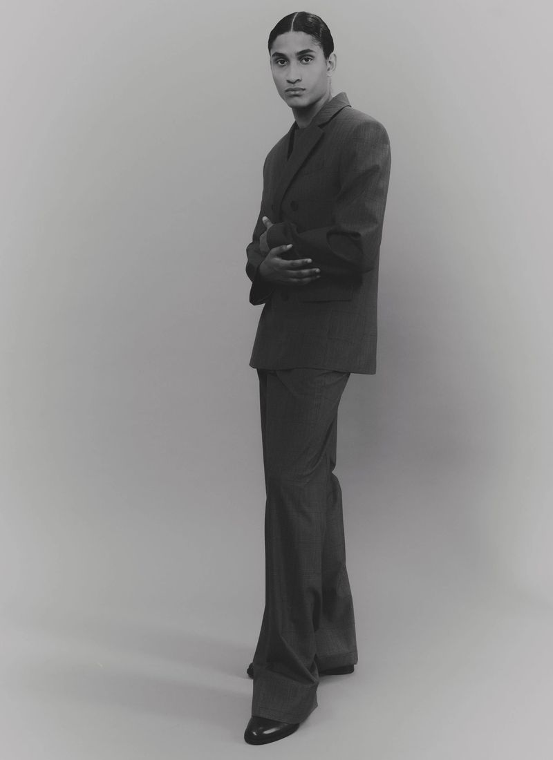 Monochromatic fashion portrait of a slender adult male in a dark suit, hands folded, against a pale background, studio lighting.