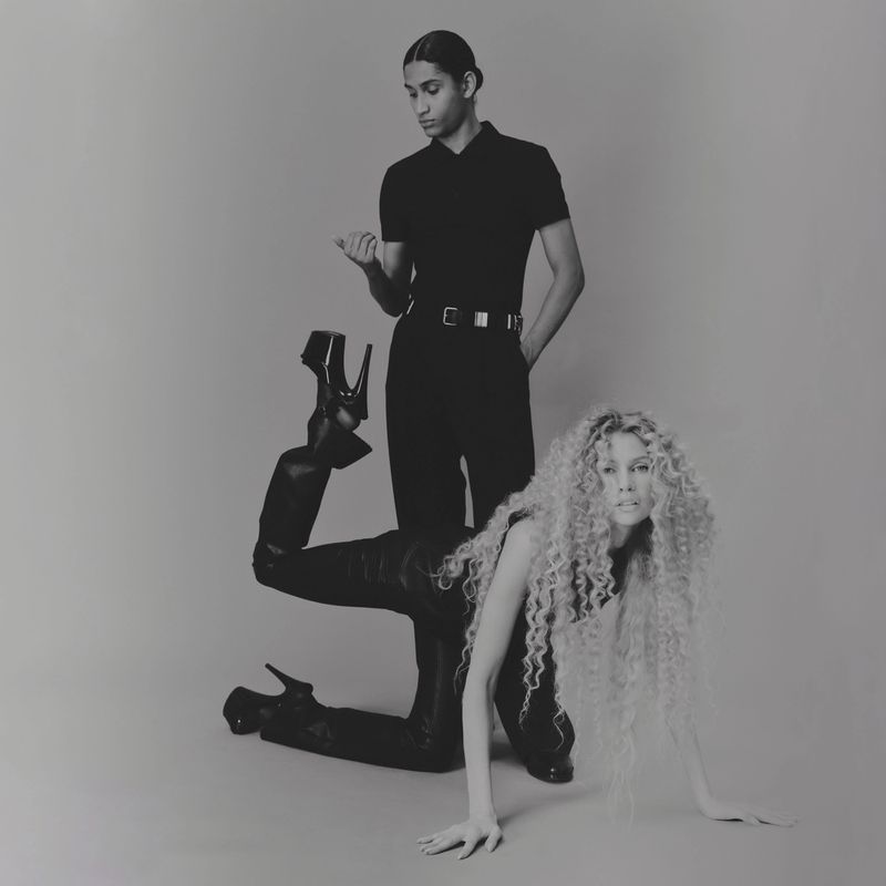 Monochrome fashion shot of a blonde woman kneeling in heels with a man standing behind her against a seamless backdrop in a studio.