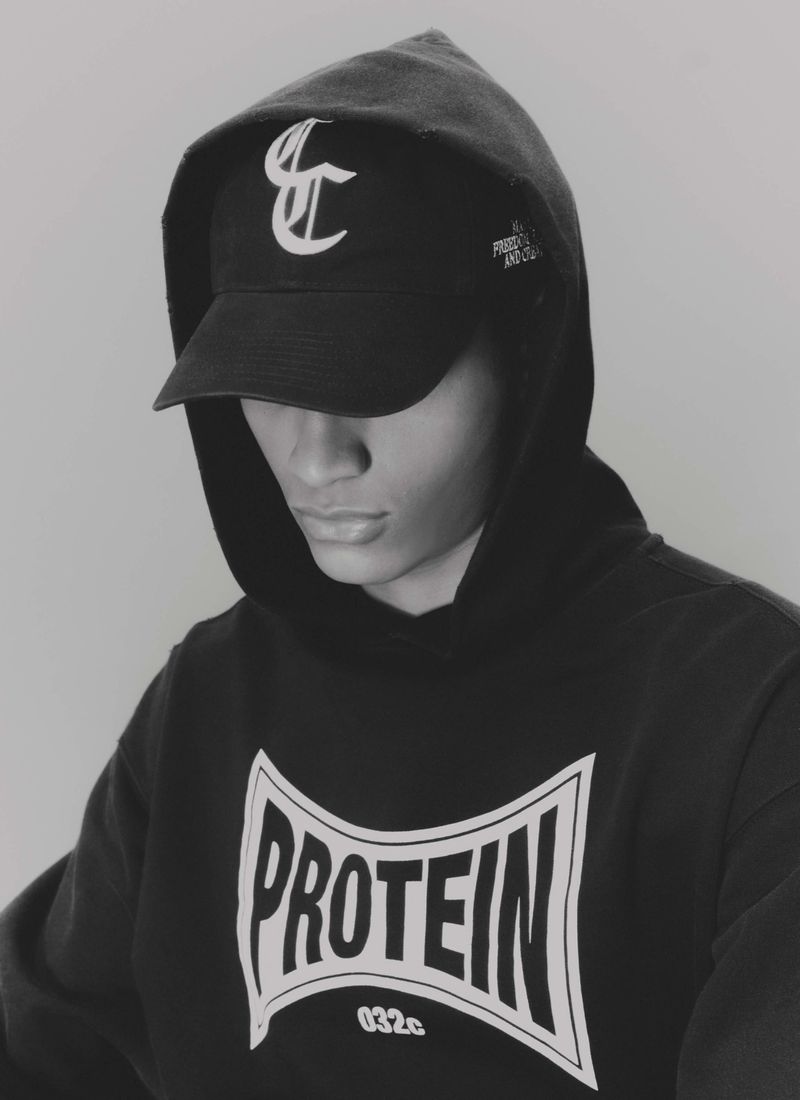 Person wearing a black hoodie with 'PROTEIN' printed on it and matching cap with a logo in a studio with a plain gray backdrop.