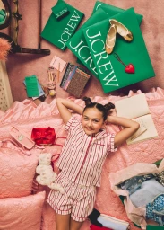 Young girl with space buns lying on pink quilted bedding surrounded by J. Crew shopping bags and gifts.