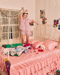 Child in pink striped pajamas jumping on a pink-dressed bed in a bedroom with patterned wallpaper for J. Crew Holiday 2025.