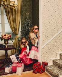 Two women holding multiple J.Crew shopping bags, peeking around a patterned wall near a Christmas tree.