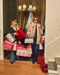 Two women carrying shopping bags and gift boxes in an interior hallway for J. Crew Holiday 2025 campaign.