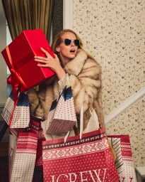 Model Abby Champion wearing sunglasses and a faux fur coat holding a large red gift box and festive shopping bags