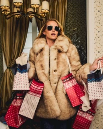 Model wearing a faux fur coat and sunglasses holding multiple holiday shopping bags indoors