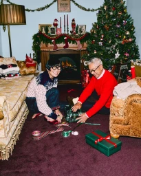 An older man and woman sitting on a dark carpet unwrapping Christmas gifts in a living room with a decorated tree.