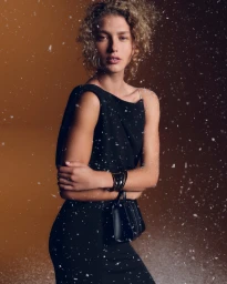 Model Dorit Revelis in a black dress wearing a bracelet against a warm backdrop with falling snow effects for Max Mara Holiday 2025.
