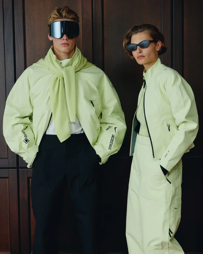Two models wearing matching lime green jackets and sunglasses standing indoors against dark wooden paneling for Nike Apres Ski 2025.