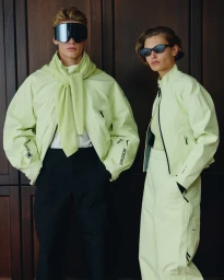 Two models wearing matching lime green jackets and sunglasses standing indoors against dark wooden paneling for Nike Apres Ski 2025.