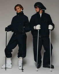 Two models wearing black ski outfits and white boots holding ski poles against a plain grey studio background.