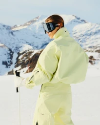 Model wearing yellow ski jacket, matching trousers, and ski goggles holding a ski pole against snowy mountain backdrop