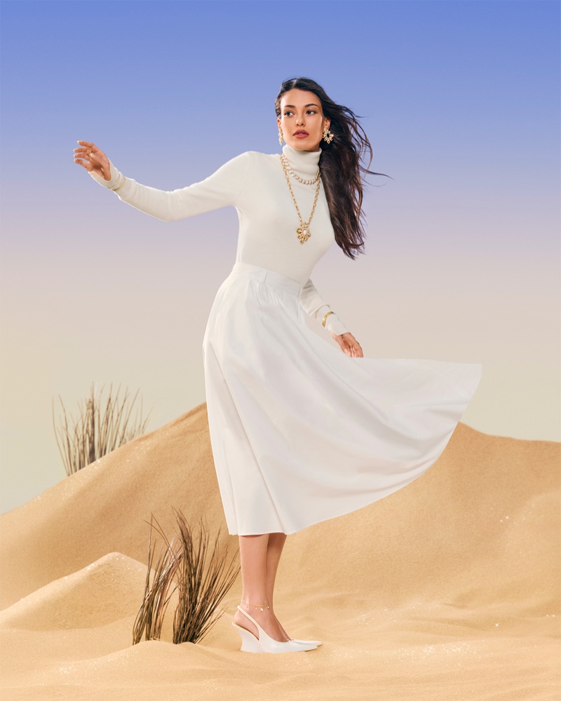 Woman wearing white turtleneck and white skirt standing on sand dunes with blue sky background