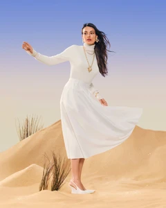 Woman wearing white turtleneck and white skirt standing on sand dunes with blue sky background