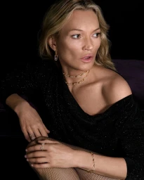 Kate Moss wearing Messika fine jewelry including gold necklaces, earrings, and rings with a black top against a dark background