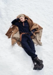 Model with blonde hair lying on a snowfield wrapped in a brown blanket wearing dark blue winter activewear and boots.