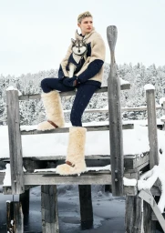 Model with short blonde hair holding a small dog while sitting on a snowy wooden dock wearing a cream and navy jacket and furry boots.