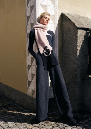 Model leaning against a stone wall wearing a long sleeve dark top, wide trousers, and a pink scarf with a round logo.