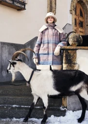 Model wearing a plaid winter coat and bucket hat standing outdoors with a goat in the foreground for Giorgio Armani