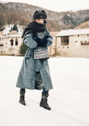 Model standing in snow wearing a long grey coat, thick scarf, winter hat, and boots with snowy buildings behind
