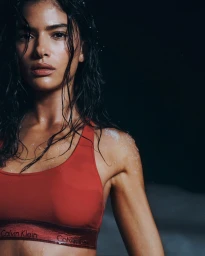 Model Rania Benchegra with wet hair wearing a red Calvin Klein sports bra against a dark background for Holiday 2025.
