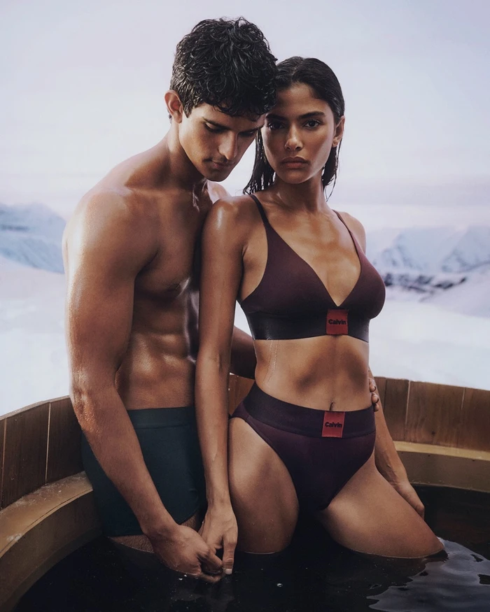 Two models standing closely together in a wooden hot tub outdoors with a snowy mountain backdrop for Calvin Klein Holiday 2025.