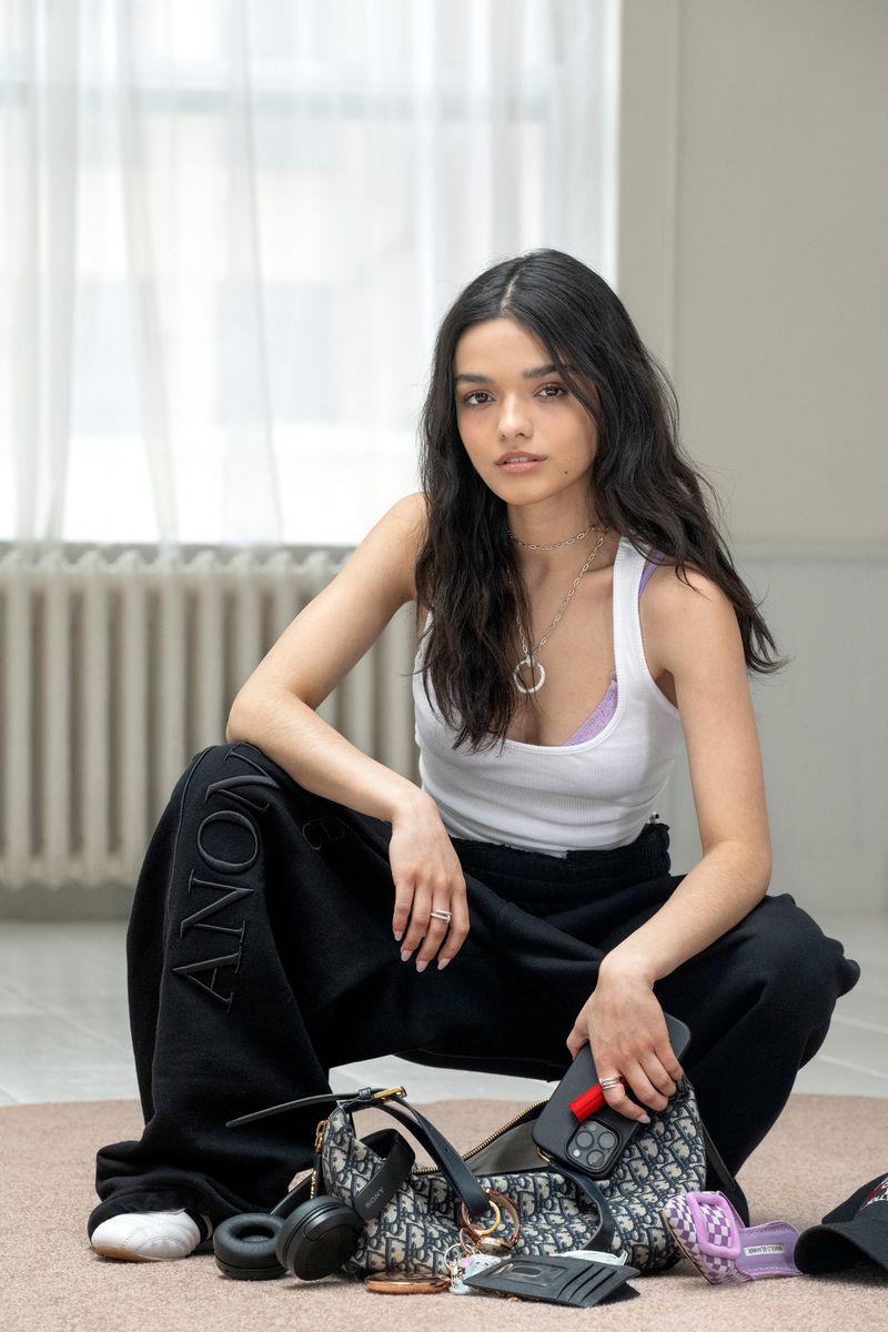 Brunette woman poses indoors wearing black sweatpants and a white tank top, kneeling behind a handbag and various personal items.