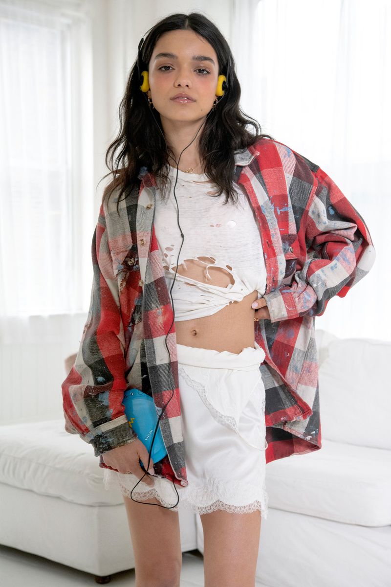 Woman in a plaid shirt and yellow headphones poses indoors with a blue portable cassette player and white ruffled shorts.