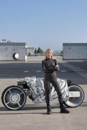 Frida Karlsson poses with arms crossed in a patterned black bodysuit next to a futuristic concept motorcycle on an open rooftop.