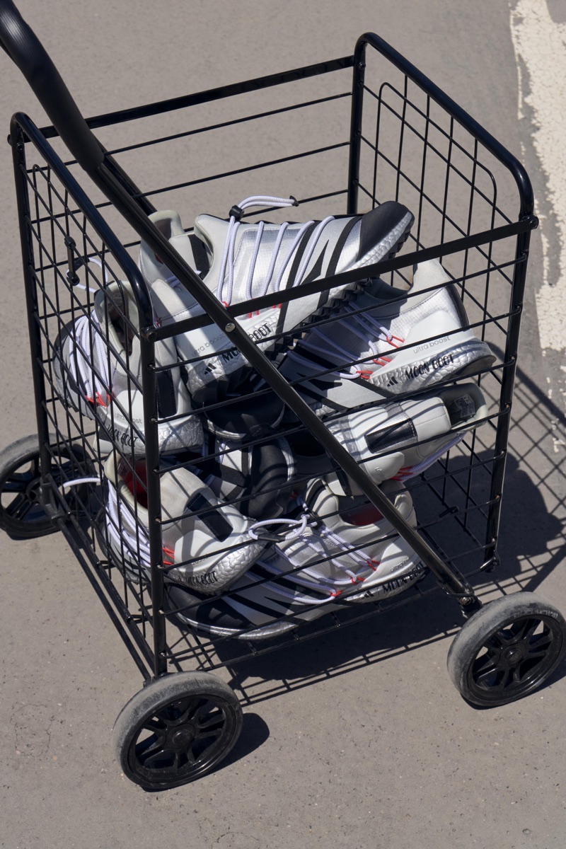 Black wire shopping cart filled with Adidas sneakers sitting on asphalt pavement outdoors.
