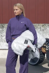 Frida Karlsson wearing a purple Adidas tracksuit and holding a metallic silver puffer jacket outdoors.