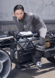 Asian woman in grey athletic bodysuit and moon boots posing on a futuristic metal vehicle frame against a concrete wall.