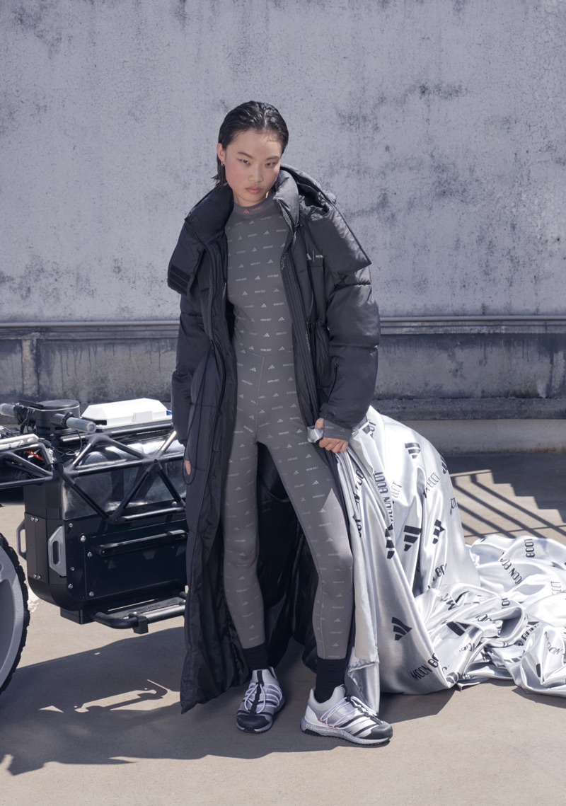 Model in long black puffer coat and grey patterned jumpsuit standing next to a vehicle against a concrete wall