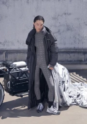 Model in long black puffer coat and grey patterned jumpsuit standing next to a vehicle against a concrete wall