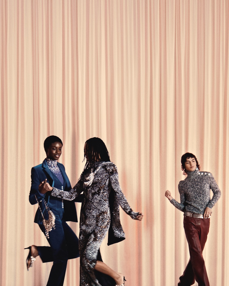 Three models in metallic and velvet eveningwear dancing in front of a pleated draped curtain backdrop.
