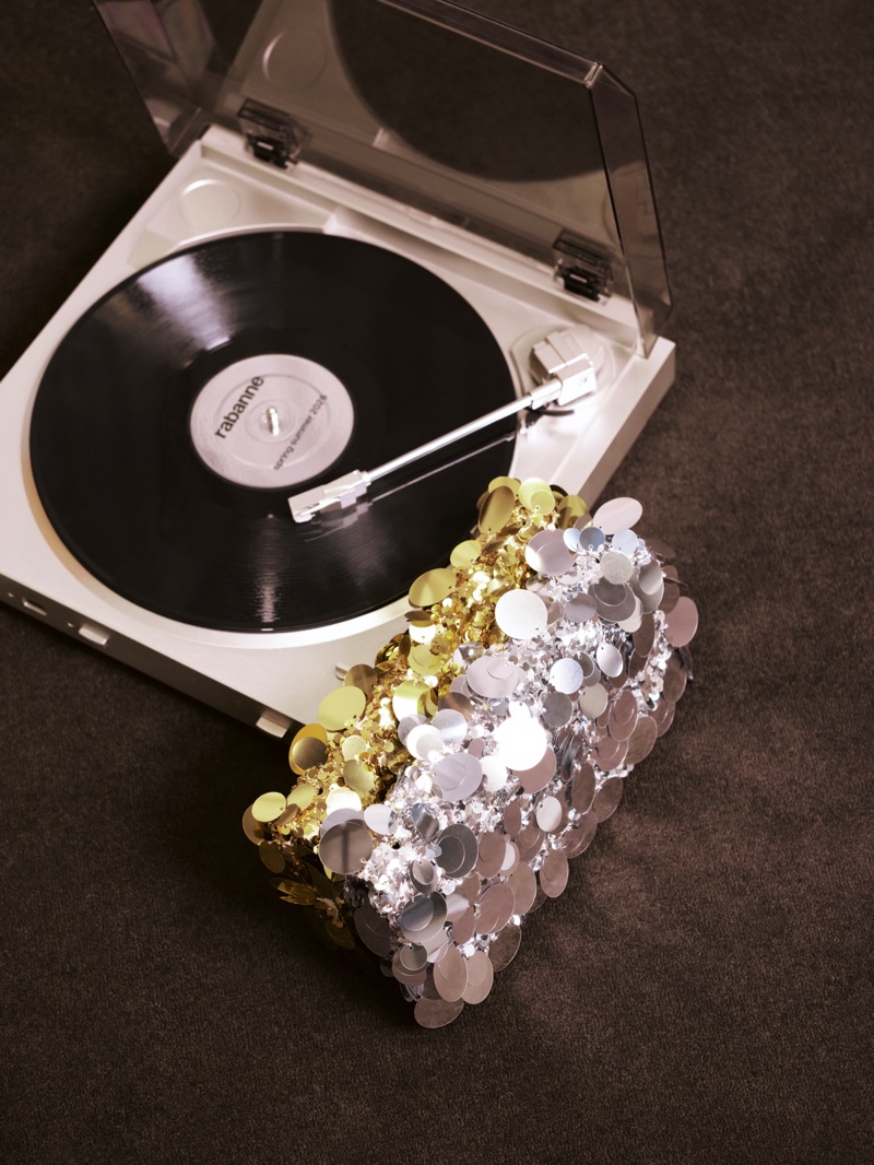 A gold and silver metallic sequin Rabanne bag rests beside a white record player with a black vinyl disc on dark carpet.
