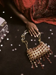 Close-up of a person wearing a red beaded dress resting a hand near a gold handbag adorned with red crystals on a dark surface.