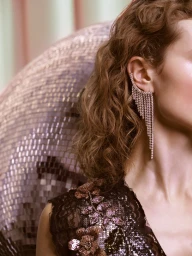 Close-up profile of a woman wearing a sparkling rhinestone fringe ear cuff and a floral embroidered top, with a disco ball background.
