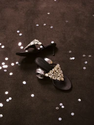 Pair of black embellished heeled sandals resting on a dark brown surface scattered with silver confetti