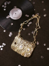 Rabanne metallic gold chainmail handbag resting on a dark surface alongside a vinyl record and scattered silver confetti.