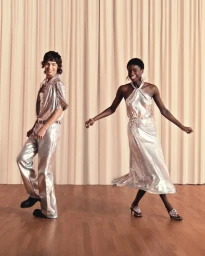 A man and a woman in metallic silver outfits dancing in front of beige curtains on a wooden floor.